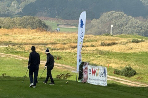 Two golfers walking together on a golf course past a WILLDOES Golf sign